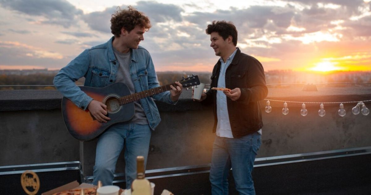 Two young men enjoy pizza and play guitar on a rooftop, embodying the carefree spirit of "your friends hunxho lyrics."